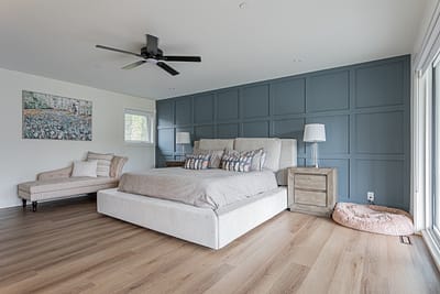 Custom additions featuring a blue paneling accent wall, large sliding doors leading to a balcony, a light gray upholstered bed, wooden nightstands, and a ceiling fan in a modern bedroom in Langley.