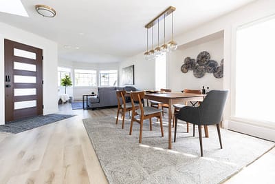 Dining room with a wooden table and six chairs, a modern chandelier, and a gray rug, connected to a living room by Bowline Construction design-build contractors in Langley, BC