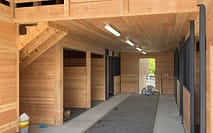 Interior of a wooden barn with stalls, a concrete aisle with a rubber mat, and a wooden loft area with railings.