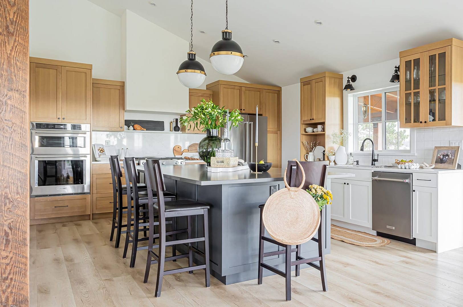 A modern kitchen with a gray island, wood cabinets, stainless steel appliances, black pendant lights, and dark wood bar stools.