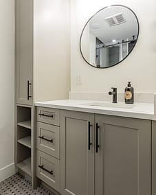 A bathroom with a glass shower enclosure, a dark vanity cabinet, a round mirror, and a wall with striped panels.