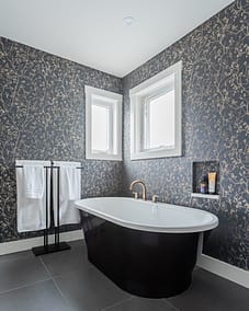 A bathroom with floral wallpaper, a black and white bathtub, two windows, and a gold faucet by home renovation contractors in Langley