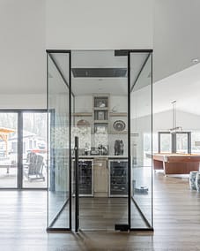 A glass enclosure with black framing in a room, containing a wine cellar, with a view of a patio and a game room