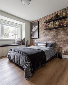 A bedroom with a bed, a brick accent wall, floating shelves, and a window seat