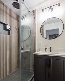 A bathroom with a glass shower enclosure, a dark vanity cabinet, a round mirror, and a wall with striped panels.