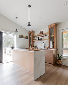 A kitchen/bar area with a light wood island, a marble countertop, wooden cabinets with glass doors, and pendant lighting