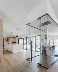 A glass enclosure with black framing in a room, containing a wine cellar, with a view of a patio and a game room