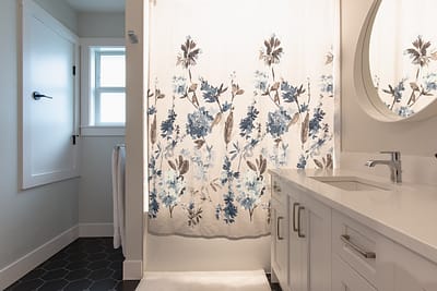 A bathroom with a floral shower curtain, white shaker cabinets, a round mirror, and a dark hexagon tile floor.