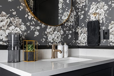 A bathroom vanity with a white quartz countertop, a gold faucet, a round mirror, and a floral wallpaper