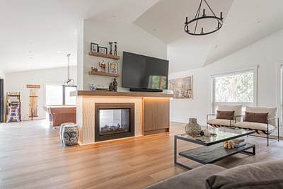 A living room with a TV mounted above a fireplace, two armchairs, a glass coffee table, and light wood floors
