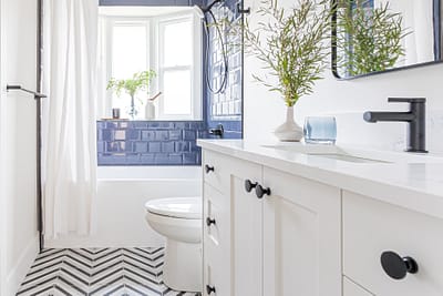 A modern bathroom with dark blue subway tile, a white vanity with a quartz countertop, black hardware, and a chevron patterned floor.