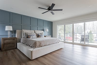Custom additions featuring a blue paneling accent wall, large sliding doors leading to a balcony, a light gray upholstered bed, wooden nightstands, and a ceiling fan in a modern bedroom in Langley.