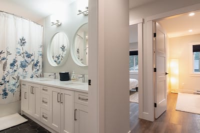 A bathroom with a double vanity, two round mirrors, a floral shower curtain, and an open doorway leading to another room.
