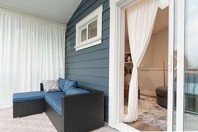 A patio featuring a dark rattan wicker sectional sofa with blue cushions, similar to a Walsunny 3-piece set, with white curtains and a sliding glass door leading indoors.