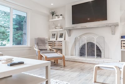 A bright living room with a white painted brick fireplace, a mounted TV, a large window, and light wood furniture custom additions in Langley, BC