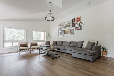 A living room with a gray sectional sofa, two armchairs, a glass coffee table, framed art on the wall, and light wood floors.