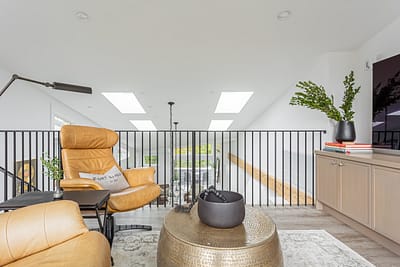 A modern loft space with a tan leather armchair featuring a pillow,a black metal railing, skylights, and a gray cabinet.