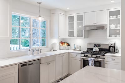 A bright white kitchen with white cabinets, stainless steel appliances, a farmhouse sink, and a large window overlooking trees by design build contractrs in Langley, BC
